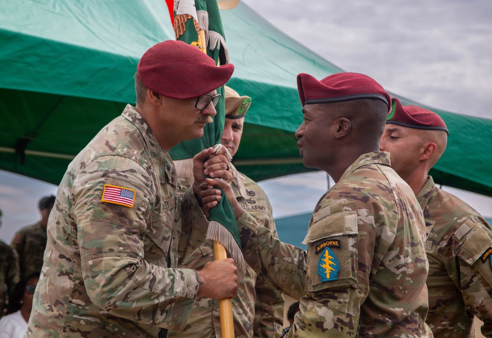 A military change of command ceremony is taking place, with two U.S. Army soldiers in maroon berets exchanging a unit guidon (flag). The soldier on the right wears an Airborne tab and a Special Forces (lightning bolt) shoulder patch, indicating this is a Special Forces unit ceremony. A green tent canopy and additional uniformed personnel are visible in the background.