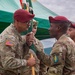 A military change of command ceremony is taking place, with two U.S. Army soldiers in maroon berets exchanging a unit guidon (flag). The soldier on the right wears an Airborne tab and a Special Forces (lightning bolt) shoulder patch, indicating this is a Special Forces unit ceremony. A green tent canopy and additional uniformed personnel are visible in the background.