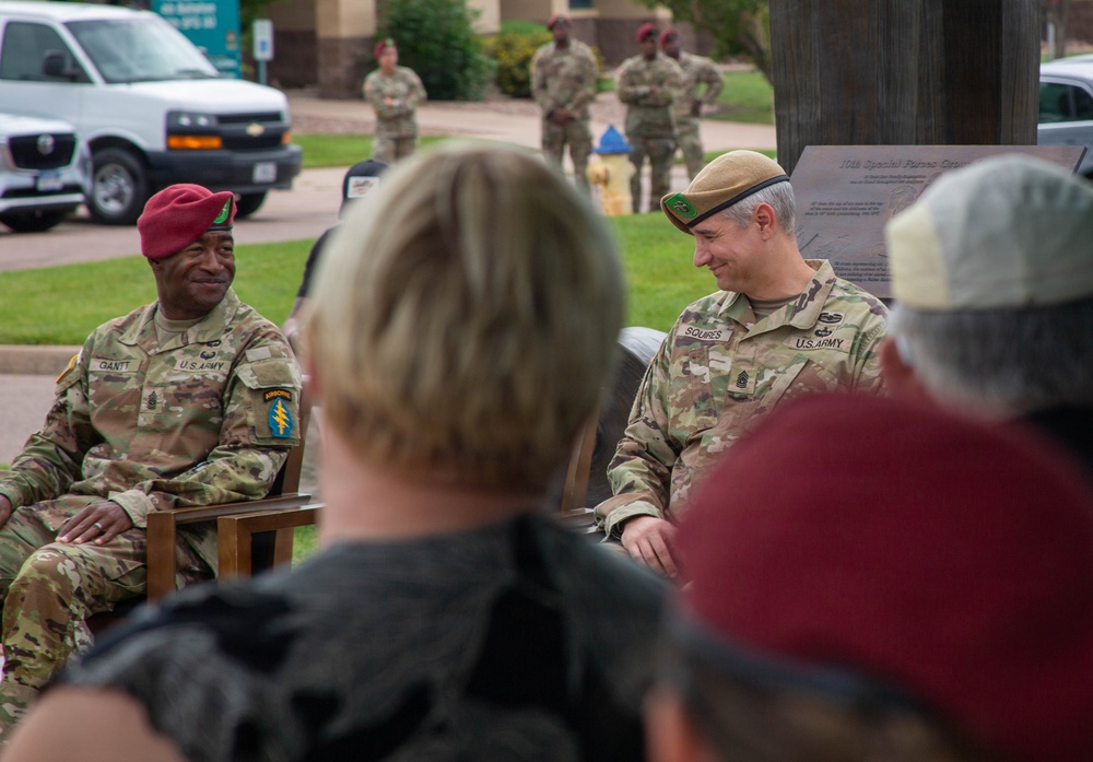 Two U.S. Army soldiers are seated at what appears to be an outdoor military ceremony or event, likely at a Special Forces installation. The soldier on the left wears a red beret and displays a Special Forces patch, while the soldier on the right wears a green beret and has the name tape 'SQUIRES' visible. Several other soldiers and civilians are present in the background near a commemorative plaque.