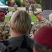 Two U.S. Army soldiers are seated at what appears to be an outdoor military ceremony or event, likely at a Special Forces installation. The soldier on the left wears a red beret and displays a Special Forces patch, while the soldier on the right wears a green beret and has the name tape 'SQUIRES' visible. Several other soldiers and civilians are present in the background near a commemorative plaque.