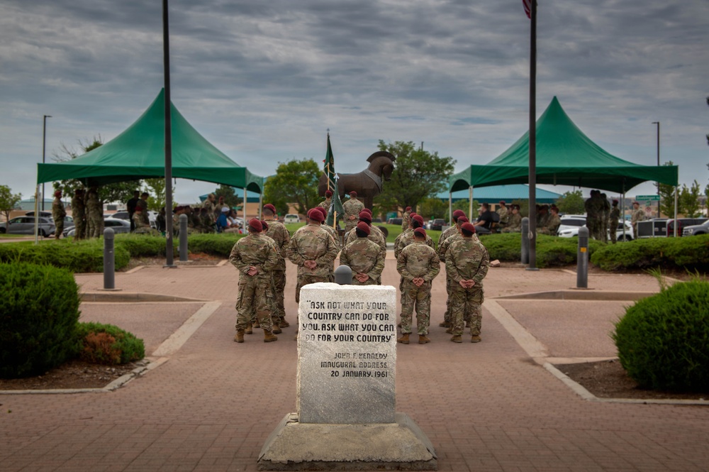 A group of military personnel wearing camouflage uniforms and red berets stand at attention in front of a stone monument inscribed with the JFK inaugural address quote. The scene appears to be a formal military ceremony at what is likely Fort Bragg or a similar U.S. Army installation, with green canopy tents set up in the background. A bronze statue and flagpoles are visible behind the assembled soldiers.