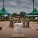 A group of military personnel wearing camouflage uniforms and red berets stand at attention in front of a stone monument inscribed with the JFK inaugural address quote. The scene appears to be a formal military ceremony at what is likely Fort Bragg or a similar U.S. Army installation, with green canopy tents set up in the background. A bronze statue and flagpoles are visible behind the assembled soldiers.