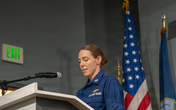 A U.S. Coast Guard officer in blue uniform is speaking at a podium during an official event. The podium displays USAA and Seattle Seahawks branding, indicating this is a military appreciation or 'Salute to Service' event. Two flags — the U.S. flag and what appears to be a Coast Guard or Navy flag — are visible in the background.