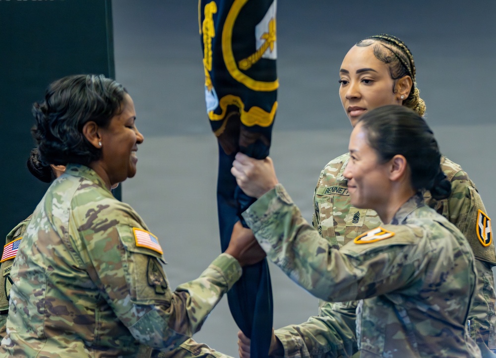 Two U.S. Army soldiers in OCP (Operational Camouflage Pattern) uniforms are engaged in what appears to be a military ceremony, likely a change of command or unit activation, involving the passing of a guidon or unit colors. A civilian woman stands in the background observing the ceremony. The soldier on the right has a visible name tape reading 'BENNETT' and both uniformed individuals wear U.S. Army shoulder patches.