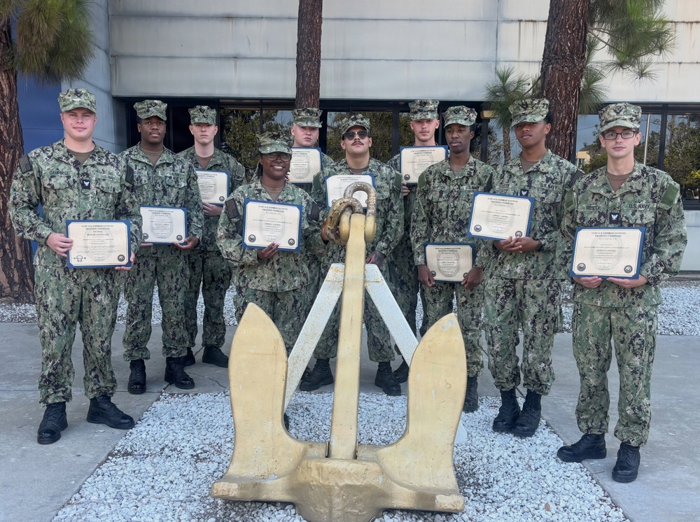 A group of approximately ten U.S. Navy personnel in Navy Working Uniform (NWU Type III) camouflage are posed together holding certificates, likely commemorating completion of a training program. They are standing in front of a large painted ship anchor displayed on a gravel bed, which serves as a symbolic backdrop. The setting appears to be an outdoor area of a naval installation or training command.