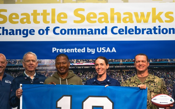Six individuals pose together at a Seattle Seahawks Change of Command Celebration presented by USAA. The group holds a blue Seahawks '12th Man' flag and a Seahawks game ball, with two participants wearing U.S. military uniforms. The event appears to honor a military change of command ceremony in partnership with the NFL team.