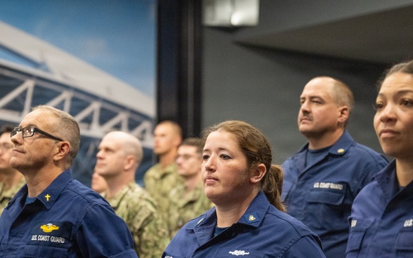 A group of U.S. Coast Guard personnel in blue operational uniforms stand at attention in an indoor facility, alongside personnel in military camouflage uniforms visible in the background. The foreground features several Coast Guard members with visible 'U.S. COAST GUARD' chest labels and various insignia. The setting appears to be a formal military ceremony or briefing room.