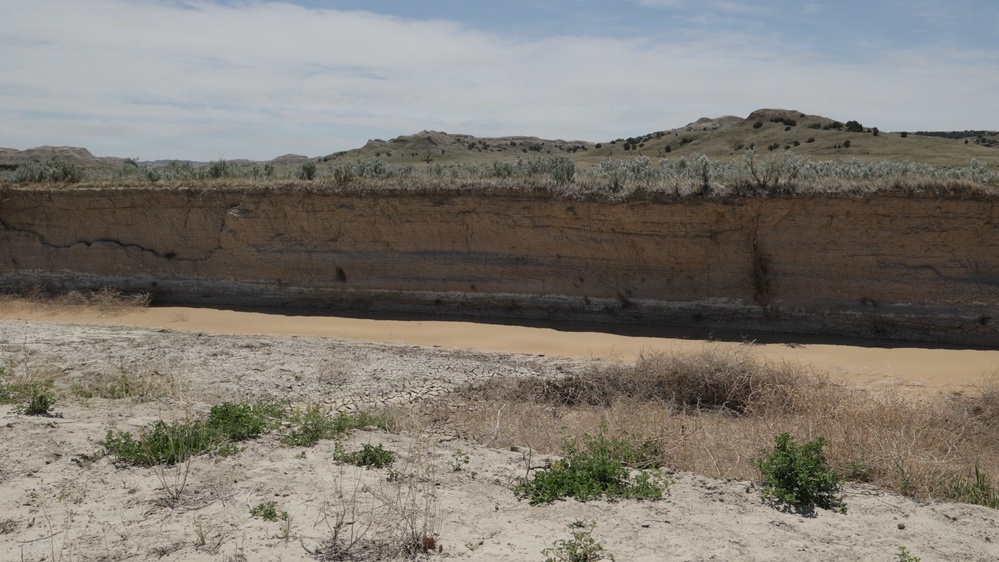 The image shows an eroded riverbank or cutbank exposing distinct sedimentary soil layers in what appears to be a semi-arid badlands environment. A shallow, sediment-laden body of water or mudflat sits at the base of the exposed cliff face, with cracked dry earth and sparse vegetation in the foreground. Rolling hills with sparse shrubs and grasses extend into the background under a partly cloudy sky.