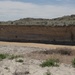 The image shows an eroded riverbank or cutbank exposing distinct sedimentary soil layers in what appears to be a semi-arid badlands environment. A shallow, sediment-laden body of water or mudflat sits at the base of the exposed cliff face, with cracked dry earth and sparse vegetation in the foreground. Rolling hills with sparse shrubs and grasses extend into the background under a partly cloudy sky.