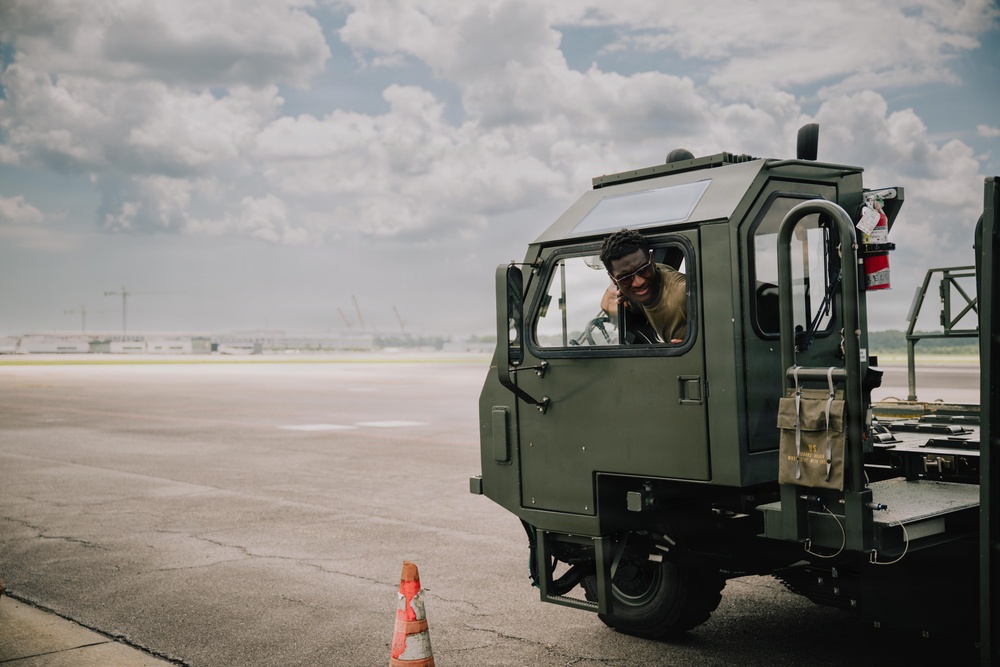 A smiling individual is seated inside the cab of a military olive-drab green ground support vehicle, leaning out of the open window. The vehicle appears to be parked on an airfield tarmac, with a traffic cone visible nearby. The scene suggests a military airfield environment during daytime with partly cloudy skies.