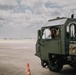 A smiling individual is seated inside the cab of a military olive-drab green ground support vehicle, leaning out of the open window. The vehicle appears to be parked on an airfield tarmac, with a traffic cone visible nearby. The scene suggests a military airfield environment during daytime with partly cloudy skies.