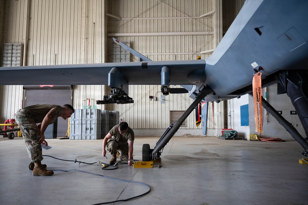 Two military personnel in camouflage uniforms are performing maintenance or pre-flight inspection on what appears to be an MQ-1 Predator or MQ-9 Reaper unmanned aerial vehicle (UAV) inside a military hangar. One technician is crouching near the landing gear while the other stands observing and appears to be taking notes. The aircraft is secured with wheel chocks and safety streamers visible on the airframe.