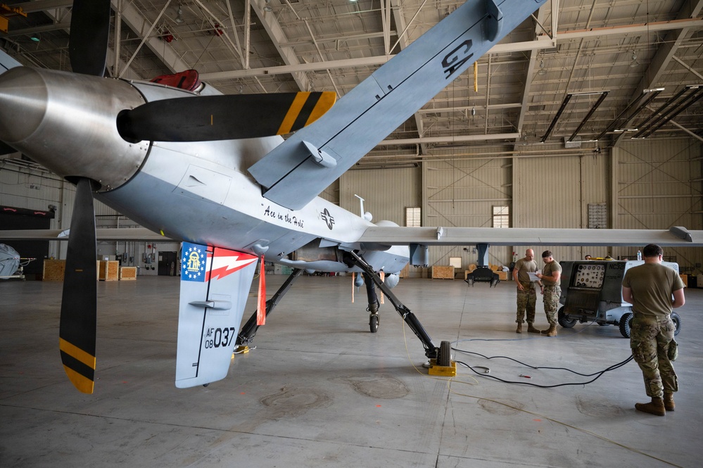 A military unmanned aerial vehicle (UAV), specifically an MQ-9 Reaper, is shown inside a large aircraft hangar. The aircraft bears the tail number 'AF 08 037' and nose art reading 'Ace in the Hole,' along with a Georgia state flag emblem on the tail. Several military personnel in camouflage uniforms are conducting maintenance or pre-flight checks near the aircraft.