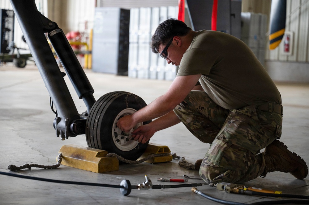A military service member in OCP (Operational Camouflage Pattern) uniform is performing maintenance on an aircraft landing gear assembly inside a hangar. The technician is kneeling on the floor and working on the wheel/tire of what appears to be a nose or main landing gear strut. Various tools and pneumatic/hydraulic lines are laid out on the floor nearby.