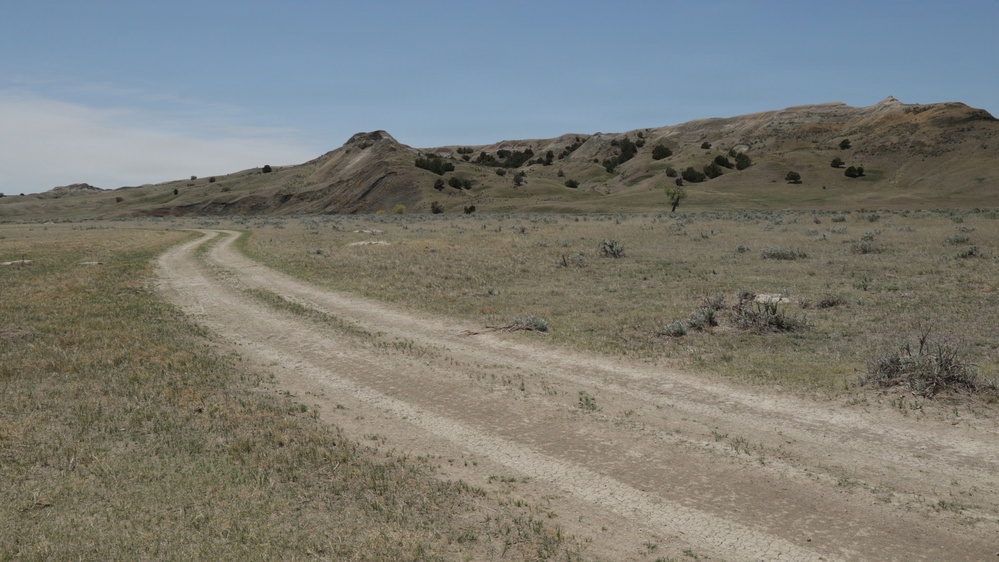 A two-track dirt road winds through an arid, semi-arid grassland landscape toward eroded buttes and hills in the background. The terrain is characteristic of the Great Plains or badlands region, featuring sparse vegetation, dry grasses, and scattered shrubs. The scene appears to be taken on a clear, sunny day with minimal cloud cover.