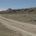 A two-track dirt road winds through an arid, semi-arid grassland landscape toward eroded buttes and hills in the background. The terrain is characteristic of the Great Plains or badlands region, featuring sparse vegetation, dry grasses, and scattered shrubs. The scene appears to be taken on a clear, sunny day with minimal cloud cover.