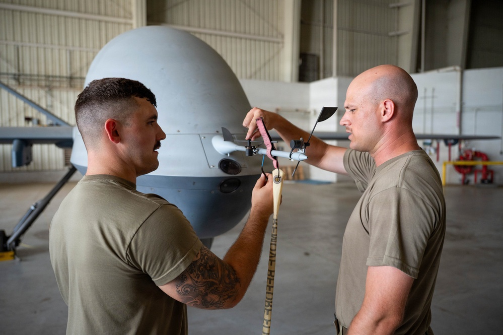 Two military personnel in olive drab t-shirts are performing maintenance or pre-flight checks on what appears to be an MQ-9 Reaper unmanned aerial vehicle (UAV) inside a hangar. One individual has a visible tattoo on his arm and is handling a strap or tether attached to the nose section of the aircraft. The setting is a military aviation hangar with the drone's distinctive bulbous nose dome prominently visible.