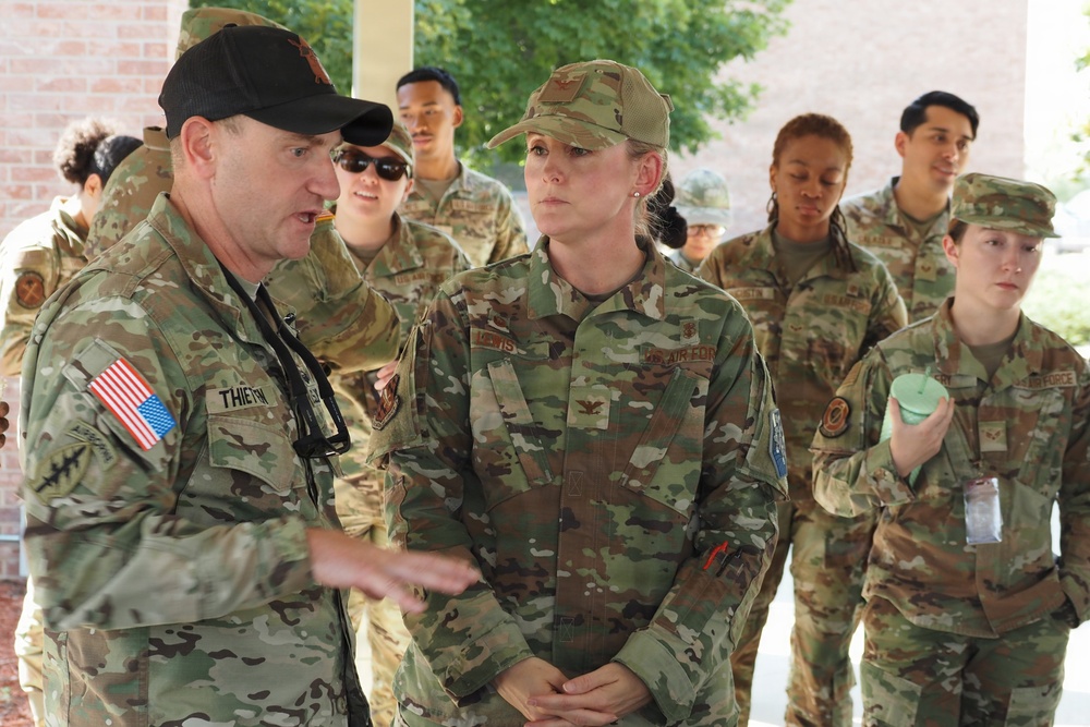 A group of military personnel in OCP (Operational Camouflage Pattern) uniforms are gathered outdoors near a brick building. A male service member wearing a black cap with a name tape reading 'THIEVIN' appears to be briefing or instructing a female service member whose name tape reads 'LEWIS' and whose uniform displays 'US AIR FORCE' tape and a colonel's eagle insignia. Several other uniformed personnel are visible in the background observing the interaction.