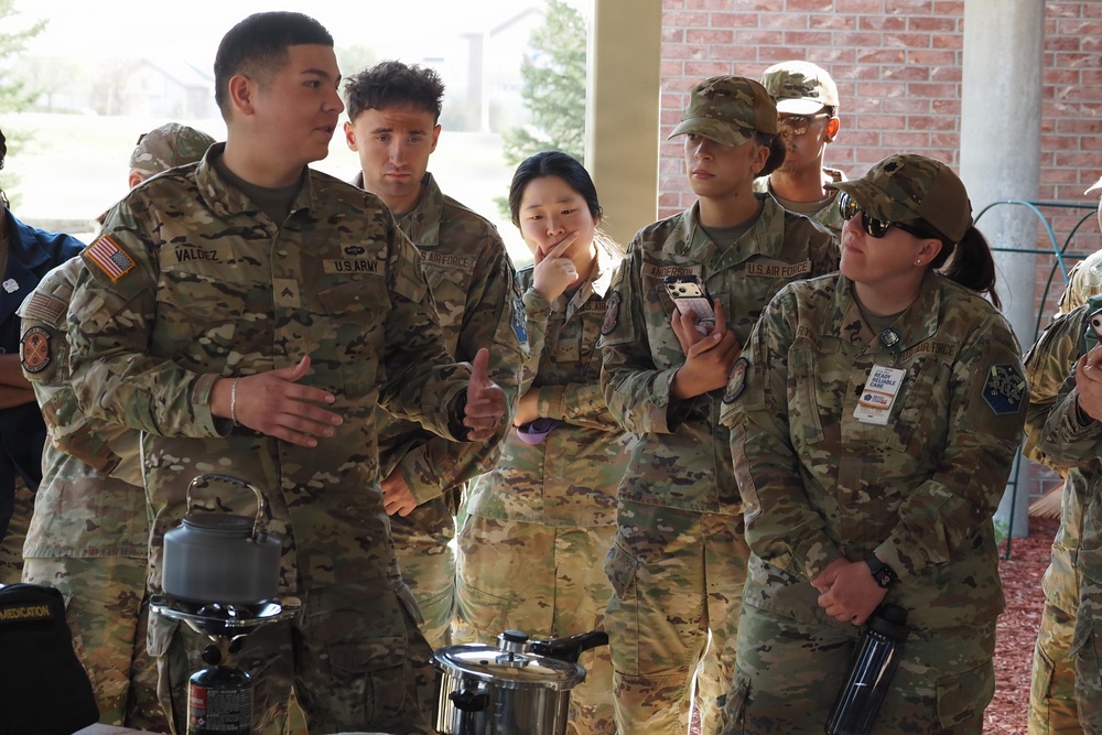 A group of U.S. military personnel in OCP (Operational Camouflage Pattern) uniforms are gathered under a covered outdoor area, appearing to receive a briefing or demonstration. The group includes both U.S. Army and U.S. Air Force members, with a soldier named 'VALDEZ' appearing to speak or instruct the others. Field equipment, including what appears to be a camp stove and a pressure cooker or canteen, is visible in the foreground.