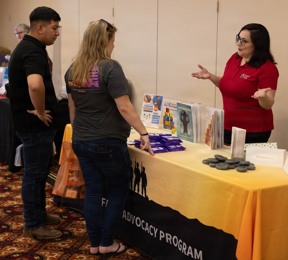 A representative in a red polo shirt is presenting information at a booth labeled 'Family Advocacy Program' during what appears to be a community fair or resource expo. Two visitors — a man in a black shirt and a woman in a gray Columbia shirt — are listening to the presenter. The table displays various pamphlets, brochures, and promotional items including wrapped snacks and small stone-like giveaways.