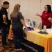A representative in a red polo shirt is presenting information at a booth labeled 'Family Advocacy Program' during what appears to be a community fair or resource expo. Two visitors — a man in a black shirt and a woman in a gray Columbia shirt — are listening to the presenter. The table displays various pamphlets, brochures, and promotional items including wrapped snacks and small stone-like giveaways.