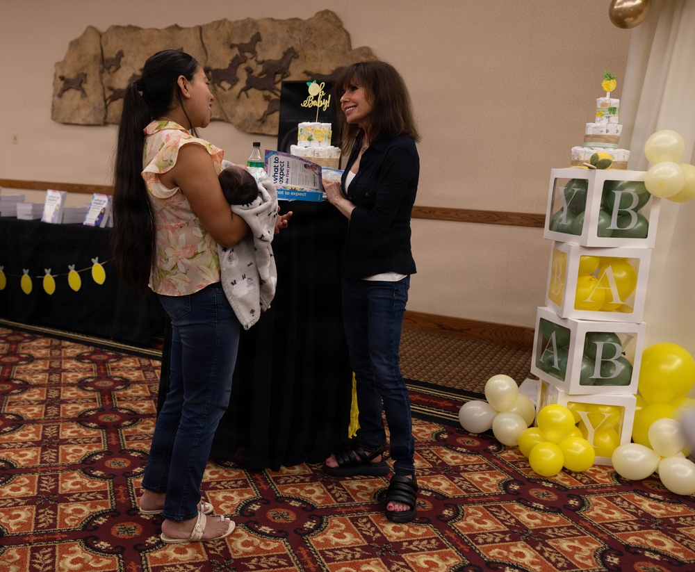 Two women are interacting at what appears to be a baby shower event held in a banquet or event hall. One woman is holding a newborn baby wrapped in a blanket while the other is presenting her with a book or informational packet. The venue is decorated with yellow and white balloons, letter blocks spelling 'BABY', a diaper cake with an 'Oh Baby!' topper, and lemon-themed garlands.