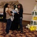 Two women are interacting at what appears to be a baby shower event held in a banquet or event hall. One woman is holding a newborn baby wrapped in a blanket while the other is presenting her with a book or informational packet. The venue is decorated with yellow and white balloons, letter blocks spelling 'BABY', a diaper cake with an 'Oh Baby!' topper, and lemon-themed garlands.