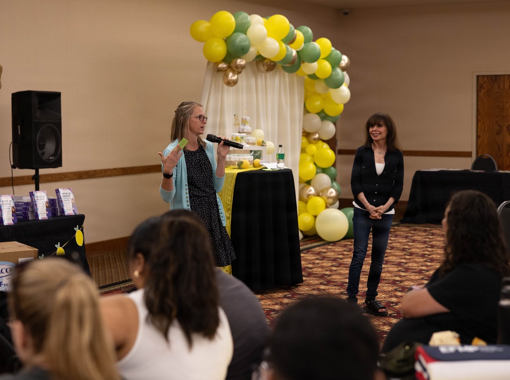 An indoor event or presentation is taking place in what appears to be a banquet or conference room. One woman is speaking into a microphone while another stands nearby smiling, with an audience seated in front of them. The space is decorated with a balloon arch in yellow, green, white, and gold colors, suggesting a celebratory or promotional gathering.