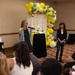 An indoor event or presentation is taking place in what appears to be a banquet or conference room. One woman is speaking into a microphone while another stands nearby smiling, with an audience seated in front of them. The space is decorated with a balloon arch in yellow, green, white, and gold colors, suggesting a celebratory or promotional gathering.