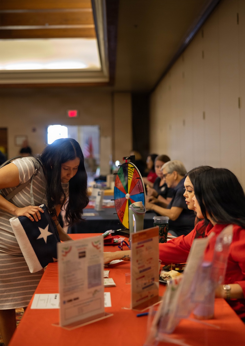 The image shows an indoor event, likely a fair, expo, or community outreach gathering, taking place in a banquet or conference hall. A woman carrying a Texas-themed tote bag leans over a red-tablecloth-covered booth table, reviewing informational materials with a seated representative dressed in red. Multiple booths and attendees are visible in the background.