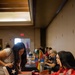 The image shows an indoor event, likely a fair, expo, or community outreach gathering, taking place in a banquet or conference hall. A woman carrying a Texas-themed tote bag leans over a red-tablecloth-covered booth table, reviewing informational materials with a seated representative dressed in red. Multiple booths and attendees are visible in the background.