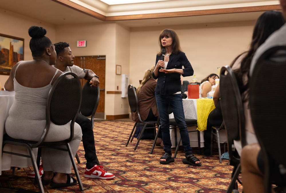 A woman stands in the center of a banquet or conference room holding a microphone, appearing to address a seated audience. The setting resembles a community meeting, workshop, or informational session held in a hotel or event hall. Attendees are seated at round tables arranged around the room.