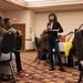 A woman stands in the center of a banquet or conference room holding a microphone, appearing to address a seated audience. The setting resembles a community meeting, workshop, or informational session held in a hotel or event hall. Attendees are seated at round tables arranged around the room.