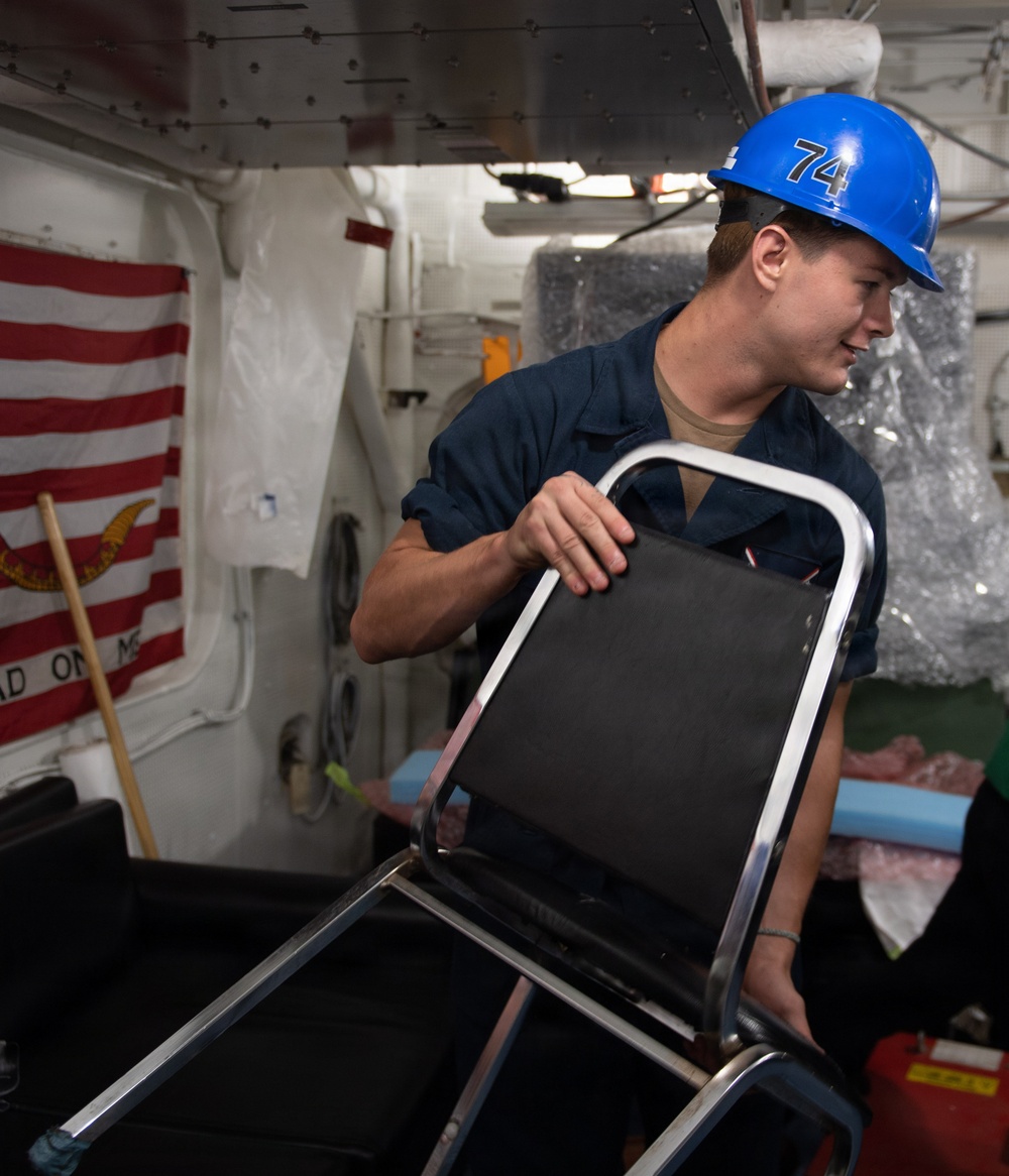 A worker wearing a blue hard hat marked with the number '74' is handling a folding chair with a chrome frame inside what appears to be a naval vessel. In the background, a 'Don't Tread on Me' (Gadsden) flag is partially visible alongside an American flag motif. The individual is dressed in a dark navy blue work uniform.