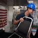 A worker wearing a blue hard hat marked with the number '74' is handling a folding chair with a chrome frame inside what appears to be a naval vessel. In the background, a 'Don't Tread on Me' (Gadsden) flag is partially visible alongside an American flag motif. The individual is dressed in a dark navy blue work uniform.