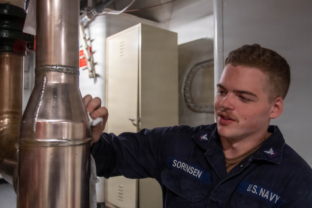 A U.S. Navy sailor identified by name tape as 'SORENSEN' is performing maintenance or cleaning on a large copper pipe aboard what appears to be a naval vessel. The sailor is wearing a dark navy blue utility uniform with a U.S. Navy insignia and an eagle rating badge visible on the chest. The setting appears to be an interior mechanical or engineering space on a ship.