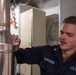 A U.S. Navy sailor identified by name tape as 'SORENSEN' is performing maintenance or cleaning on a large copper pipe aboard what appears to be a naval vessel. The sailor is wearing a dark navy blue utility uniform with a U.S. Navy insignia and an eagle rating badge visible on the chest. The setting appears to be an interior mechanical or engineering space on a ship.