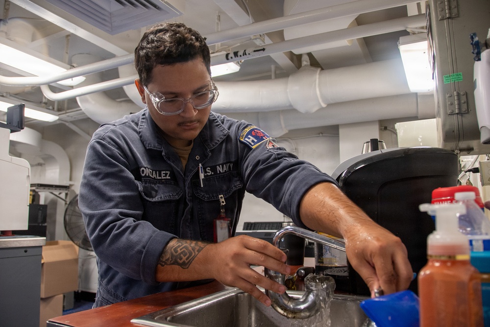 A U.S. Navy sailor named Gonzalez, wearing a blue Navy coverall uniform and safety glasses, is working at a sink in what appears to be an interior compartment of a naval vessel. He appears to be performing maintenance or repair work on a faucet/sink fixture. The environment shows typical shipboard interior features including exposed pipes, overhead lighting, and industrial equipment.