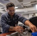 A U.S. Navy sailor named Gonzalez, wearing a blue Navy coverall uniform and safety glasses, is working at a sink in what appears to be an interior compartment of a naval vessel. He appears to be performing maintenance or repair work on a faucet/sink fixture. The environment shows typical shipboard interior features including exposed pipes, overhead lighting, and industrial equipment.