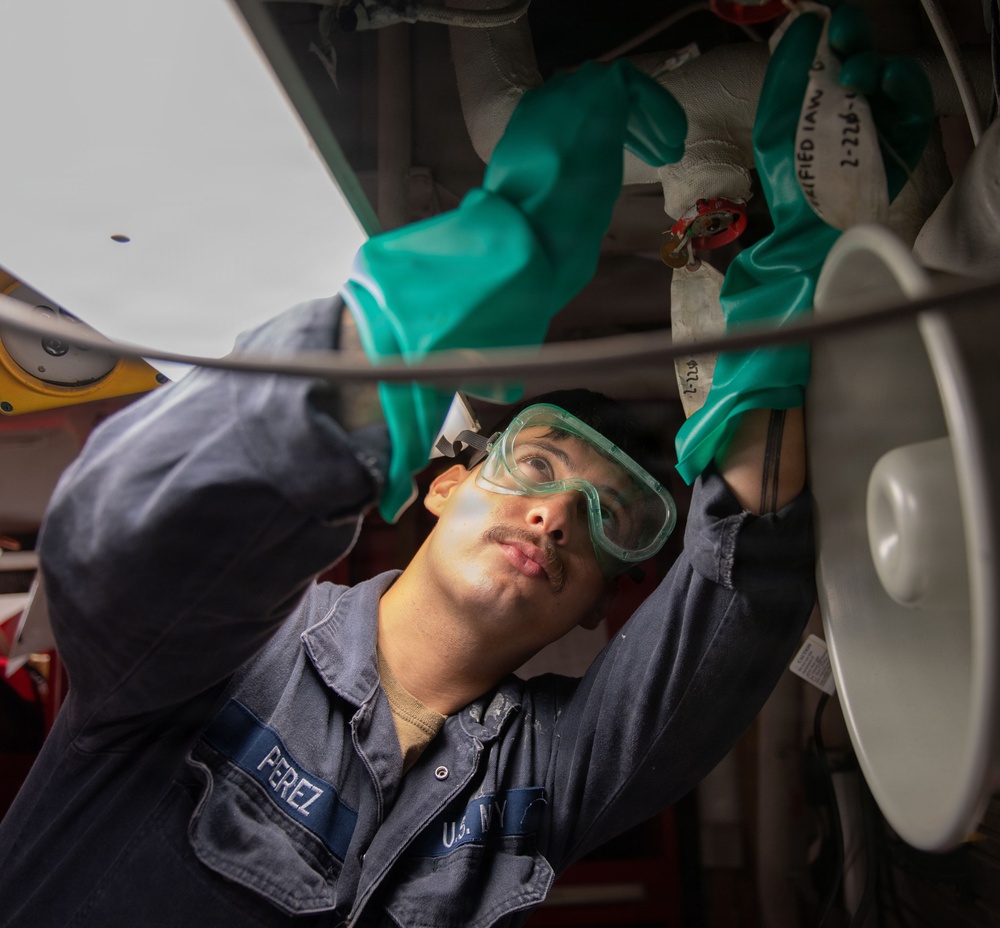 A U.S. Navy sailor named Perez is performing maintenance work on what appears to be aircraft or naval equipment. The sailor is wearing protective gear including green nitrile gloves and safety goggles while working overhead on mechanical components. The image is taken from a low angle, looking up at the worker as they inspect or service the underside of the equipment.