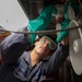 A U.S. Navy sailor named Perez is performing maintenance work on what appears to be aircraft or naval equipment. The sailor is wearing protective gear including green nitrile gloves and safety goggles while working overhead on mechanical components. The image is taken from a low angle, looking up at the worker as they inspect or service the underside of the equipment.