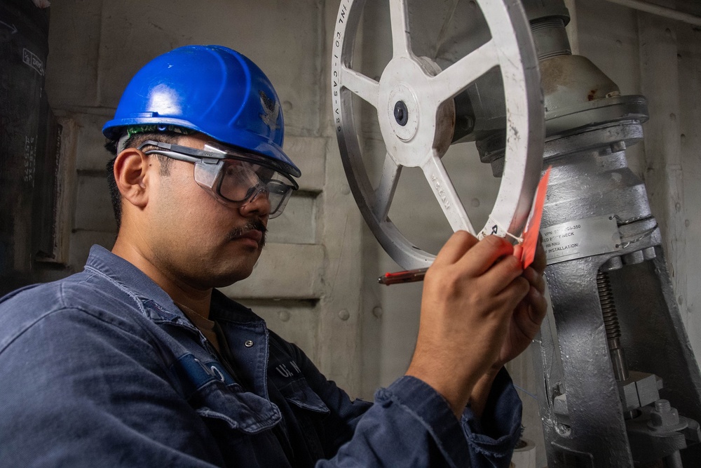 A worker wearing a blue hard hat and safety goggles is inspecting or labeling a large manual valve handwheel in an industrial or naval engineering space. The worker appears to be attaching or writing on a red tag affixed to the white-painted handwheel. The setting suggests a ship's engineering compartment or industrial facility based on the visible piping, valve hardware, and instructional labels.