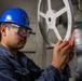 A worker wearing a blue hard hat and safety goggles is inspecting or labeling a large manual valve handwheel in an industrial or naval engineering space. The worker appears to be attaching or writing on a red tag affixed to the white-painted handwheel. The setting suggests a ship's engineering compartment or industrial facility based on the visible piping, valve hardware, and instructional labels.