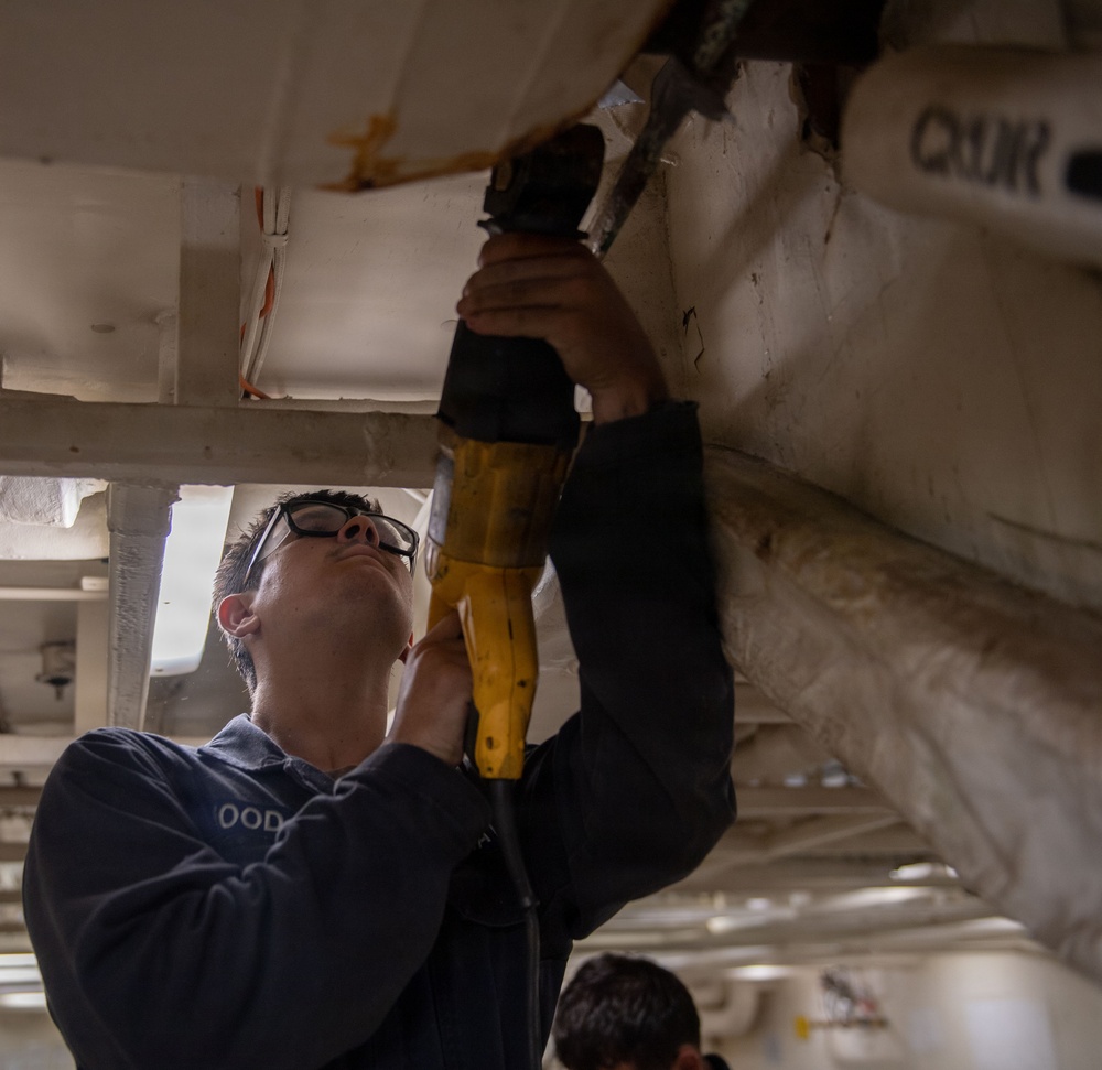A worker wearing safety glasses and a dark uniform with 'OOD' visible on the chest is using a yellow power tool (reciprocating saw or drill) to work on an overhead structure. The work appears to involve cutting or drilling into a white-painted surface, possibly part of a ship or large vehicle interior. A second person is partially visible in the background.
