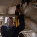 A worker wearing safety glasses and a dark uniform with 'OOD' visible on the chest is using a yellow power tool (reciprocating saw or drill) to work on an overhead structure. The work appears to involve cutting or drilling into a white-painted surface, possibly part of a ship or large vehicle interior. A second person is partially visible in the background.
