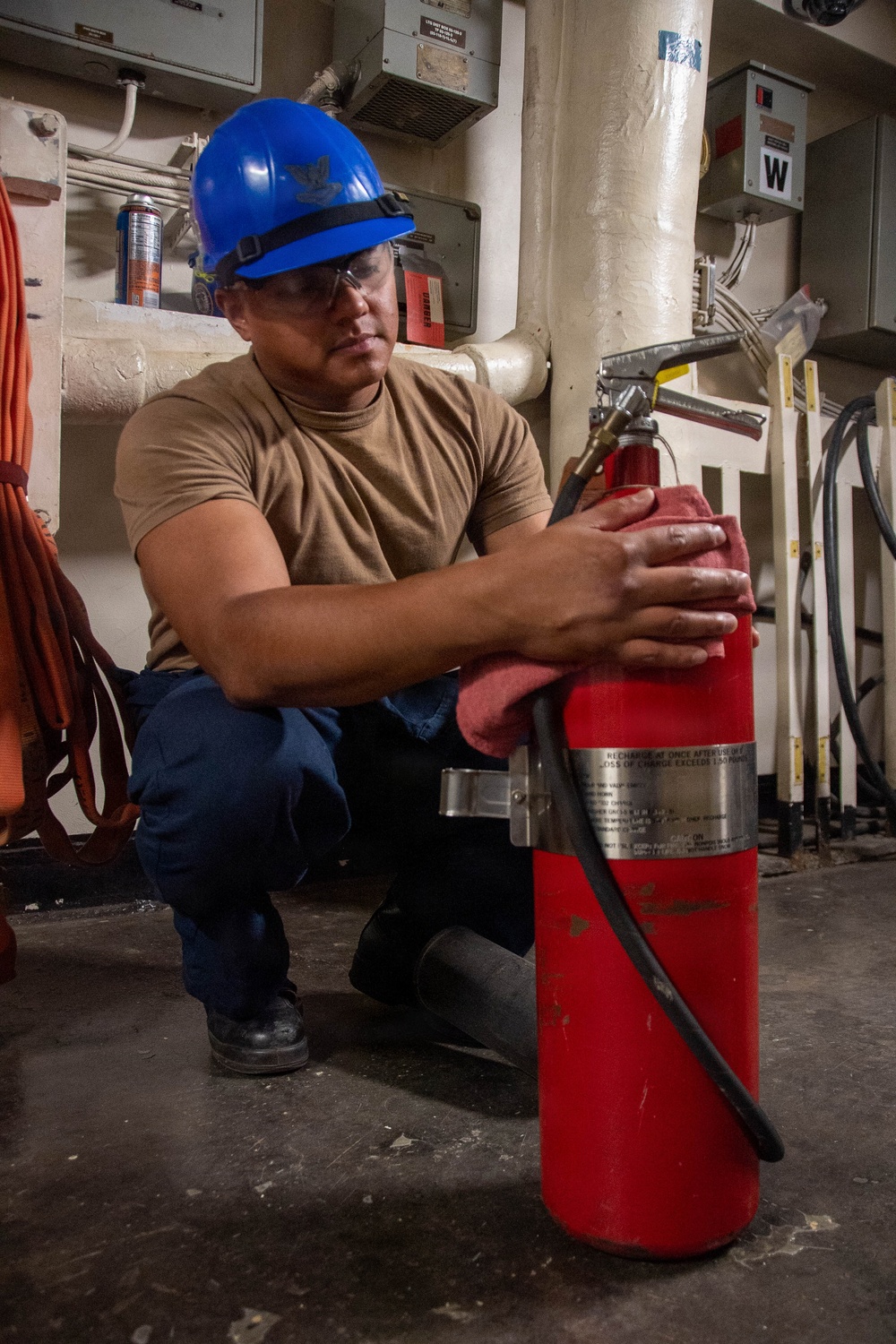 A worker wearing a blue hard hat with an eagle emblem and safety glasses is crouching down while cleaning or inspecting a large red fire extinguisher using a red cloth. The setting appears to be an industrial or naval facility interior, with pipes, electrical panels, and cables visible in the background. The activity suggests routine maintenance or inspection of fire safety equipment.