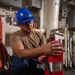 A worker wearing a blue hard hat with an eagle emblem and safety glasses is crouching down while cleaning or inspecting a large red fire extinguisher using a red cloth. The setting appears to be an industrial or naval facility interior, with pipes, electrical panels, and cables visible in the background. The activity suggests routine maintenance or inspection of fire safety equipment.