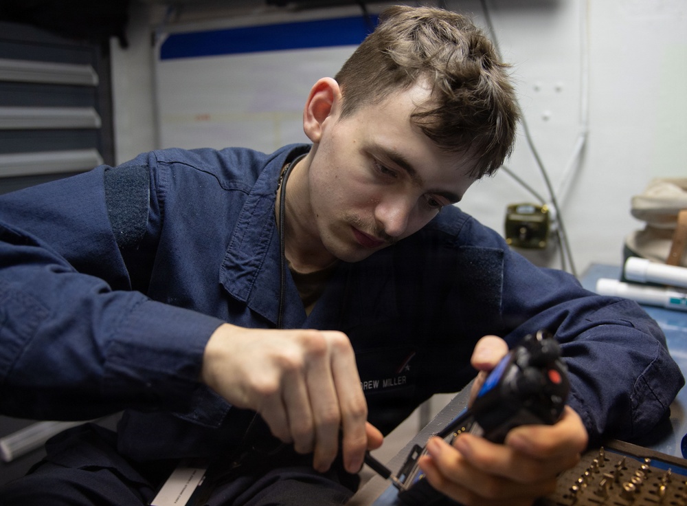 A young male service member in navy blue coveralls is focused on performing maintenance or assembly work on a firearm, likely a pistol. He is working at a workbench in what appears to be a military workspace or armory. Small brass components, possibly cartridges or parts, are visible on the work surface in front of him.