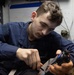 A young male service member in navy blue coveralls is focused on performing maintenance or assembly work on a firearm, likely a pistol. He is working at a workbench in what appears to be a military workspace or armory. Small brass components, possibly cartridges or parts, are visible on the work surface in front of him.
