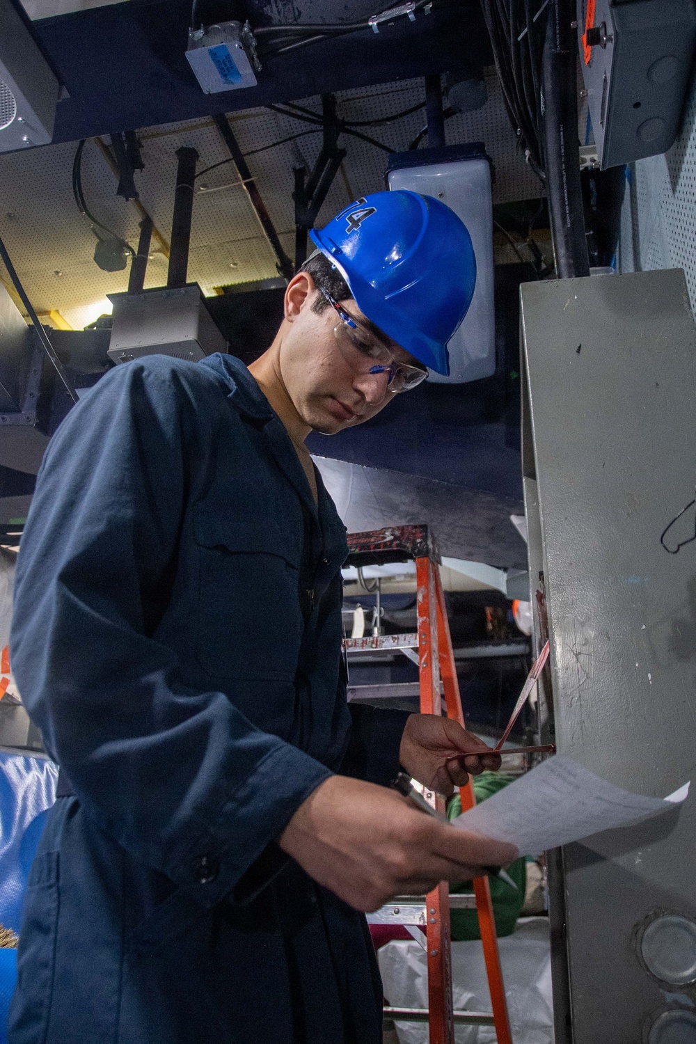 A young male technician or worker wearing a blue hard hat and safety glasses is examining a document or checklist while standing next to a metal electrical or equipment panel. He is dressed in dark navy blue coveralls and appears to be in an industrial or mechanical workspace. An orange ladder is visible in the background among various cables, equipment, and overhead infrastructure.